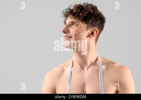 Young man with markings on his face and tape measure against grey background, closeup. Plastic surgery concept Stock Photo