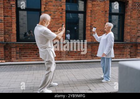 Wide shot of happy senior couple capturing moments in city street with smartphone on sunny summer day, enjoying retirement, exploring urban landscapes Stock Photo