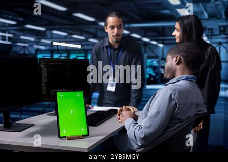 Multiethnic engineering team works on green screen tablet in data center housing storage gear. Diverse staff members using mockup device in server hub with networking components Stock Photo