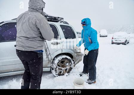 Travel in wintertime, pair of friends, man and woman, putting chains on the wheels of an SUV vehicle, in order to be able to move forward over the sno Stock Photo