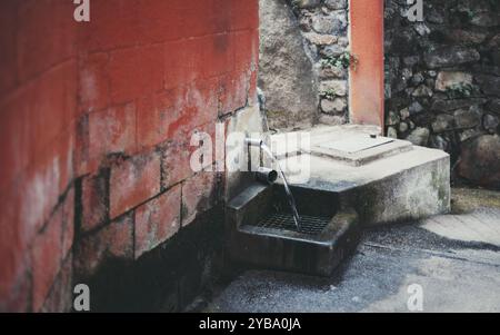 Outdoor water fountain built into a red, weathered wall with mossy bricks. A metal spout pours water into a rectangular basin with a metal grate, surr Stock Photo