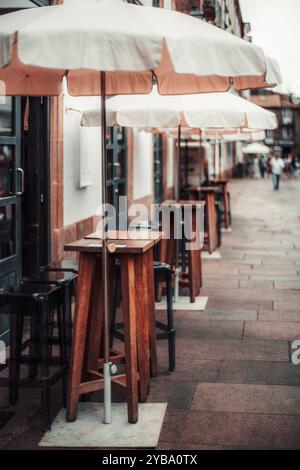 Outdoor cafe with wooden high tables, bar stools, and white umbrellas lining a city street. Shallow depth of field emphasizes the distant street. Sele Stock Photo