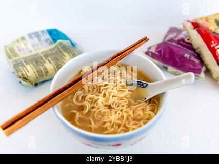 A bowl of instant noodles in soup base with a pair of chopsticks and ceramic spoon, and packets of the noodles isolated on white. Stock Photo