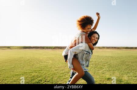 Two young women enjoying a victorious piggyback ride in a sunny field, capturing the essence of freedom, friendship, and carefree fun in the great out Stock Photo