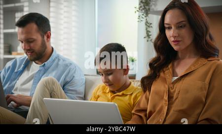 Caucasian family man woman father mother parents working remote online on laptop kid boy child son playing on mobile phone smartphone on couch home Stock Photo