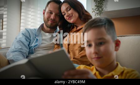 Smiling kid boy child writing notes in notebook drawing do homework at home loving parent mother father mom dad sitting in embrace on couch looking Stock Photo