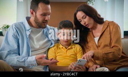 Happy family father mother parents son kid boy child talk communicate looking smartphone screen together on couch home using mobile app for play watch Stock Photo
