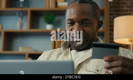 Happy african american man holding credit card using instant easy payment on laptop at home remote making purchase in online store smiling male Stock Photo