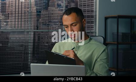 African american man manager looking at laptop screen writing financial report analyzing data from internet young ethnic businessman write notes doing Stock Photo