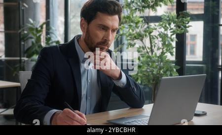 Pensive puzzled Caucasian business man businessman male entrepreneur corporate employee sales manager working in office typing on laptop searching Stock Photo