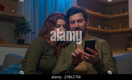 Happy family Caucasian married couple husband wife man woman shopping in online store on cozy couch at home evening using mobile application on phone Stock Photo