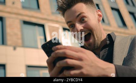 Enthusiastic happy surprised man reading good news using mobile phone outdoors in sunlight lucky satisfied male businessman winner win online bet Stock Photo