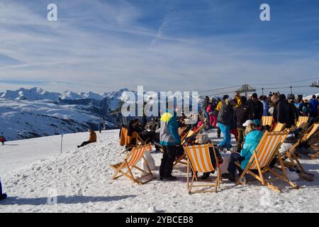 Les Menuires - March 13 2024: Apres ski party by an active ski slope in Les Menuires French ski resort. Skiers drinking next to snowcapped mountains. Stock Photo