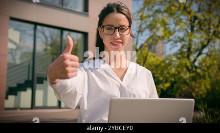 Smiling Caucasian woman girl female businesswoman freelancer student using laptop in city outdoors showing thumbs up gesture approval recommending Stock Photo