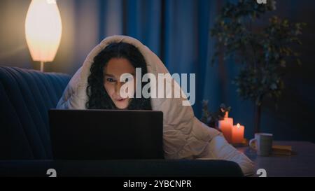 Hispanic freelancer girl Latino woman Indian Arabian female at night evening home at couch sofa under blanket cover duvet working with laptop watching Stock Photo