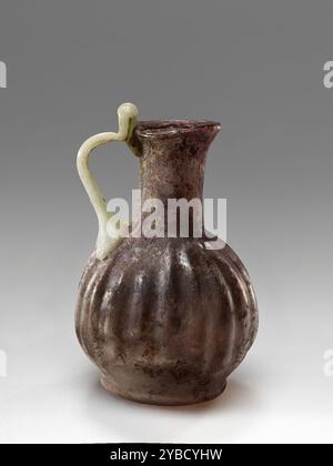 Oinochoe, 1st-2nd century A.D.  Additional Info: This mold-blown oinochoe is made of purple glass. The seam mark indicates that this oinochoe was blown in a two-part mold. Its spherical ribbed body rests on a flat base. The cylindrical neck ends with a flaring mouth and lip folded inwards. An opaque white handle with an elbow has been applied on the shoulder and drawn up to the lip. The surface presents some iridescence on the inside as well as dark accretions and discoloration. A small fill has been added on the rim. Stock Photo