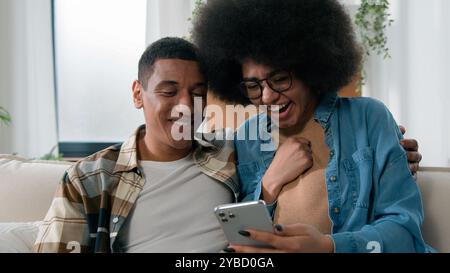 African American happy couple having fun using smartphone excited browsing mobile phone app at home girl and guy man and woman talking shopping Stock Photo