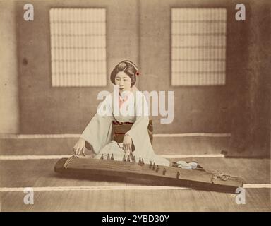 Japanese Woman Playing a Koto, 1865-1875. A Japanese woman wearing kimono and seated on the floor, playing a koto. Her fingers are posed over the strings of the koto. Stock Photo