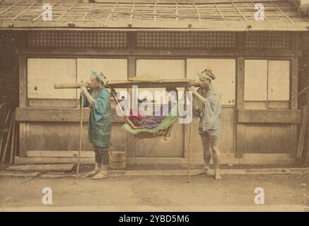 Japanese woman being carried by two Japanese men in a kago, 1868-1880. View of a woman sitting in a kago, or palanquin, being held by two men who each also hold a tall walking pole. They stand on a dirt road in front of a house. Stock Photo