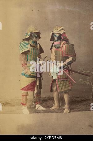 Two Japanese warriors in full dress costumes, 1868-1880. Studio portrait of two samurai in full armor. The two men stand facing each other, each resting one hand on a sword strapped to his waist. Grass beneath their feet and a small tree act as props. Stock Photo