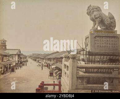 Gionmachi, Kioto, about 1882-1890s. Looking westward along the busy Shijo Street (Shijo-dori) from the western gate of Yasaka Shrine, also known as Gion Shrine. Visible in the foreground is one of the two guardian dog statues that flank the steps of the gate. A watchtower can be seen on the center left area of the image. Stock Photo