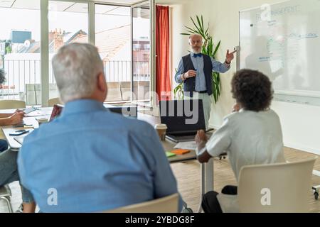 Elderly bearded doctor explaining pain management techniques to diverse medical team. Professional healthcare seminar in bright office setting with wh Stock Photo