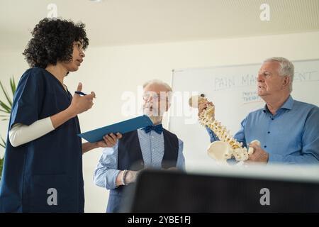 Diverse group of medical professionals collaborating on pain management strategies. Young doctor presents to senior colleagues while spine model is de Stock Photo
