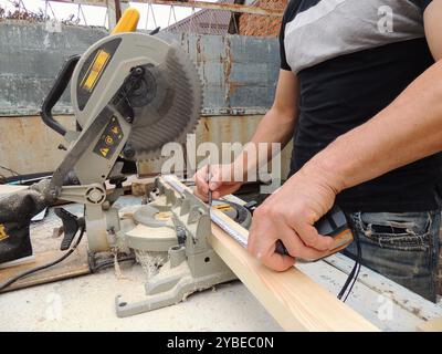 a carpenter measures the required size on a wooden plank with a tape measure on a workbench with a standing end saw, taking measurements Stock Photo
