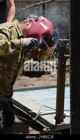 welder in red protective mask welds metal pipe to iron leg of inverted table, welding of iron parts in metal furniture manufacturing process Stock Photo