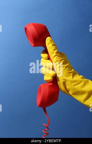 Woman in rubber glove holding red telephone handset on blue background, closeup Stock Photo