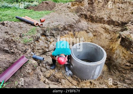 Using rotary hammer with chisel, worker punches hole for sewer pipe in concrete septic tank ring. Stock Photo