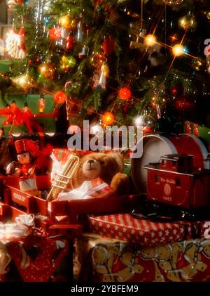 A collection of wrapped presents sits under a Christmas tree decorated with ornaments and lights. A red toy train, a teddy bear with a trumpet, and a Stock Photo