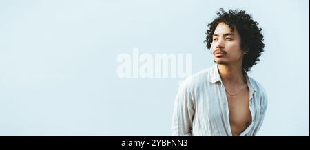 Portrait of a young asian man with curly hair, wearing an open, light linen shirt, looking thoughtfully to the side against a clean, soft blue backgro Stock Photo