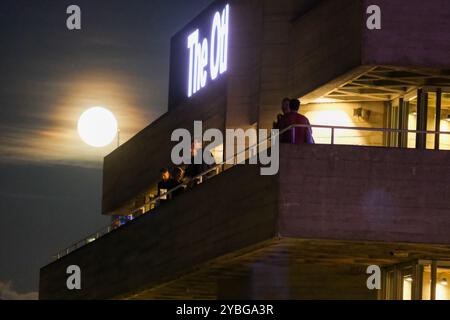 London, UK. 18th October 2024. Patrons at the National Theatre enjoy a moonlit scene during a performance interval as a waning Hunter's supermoon rises on Friday evening against the City skyline. A supermoon occurs when the earth's satellite is closer than usual, and this month is the brightest of the four scheduled this year. Credit: Eleventh Hour Photography/Ģ����ý Live News Stock Photo