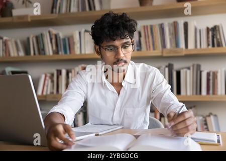 Focused smart male Indian student reading textbook in library Stock Photo