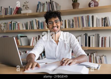 Cheerful young Indian college student in eyeglasses sitting at table Stock Photo
