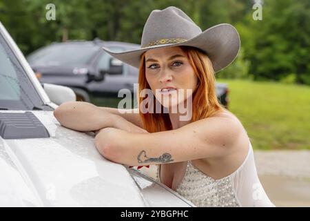 A beautiful redhead model poses in a country setting wearing cowgirl attire against her truck Stock Photo