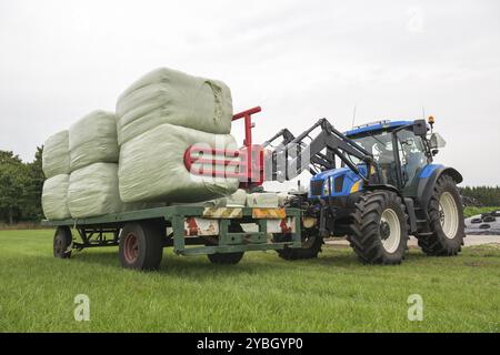 Loading plastic hay bales on a flat cart by a blue tractor Stock Photo