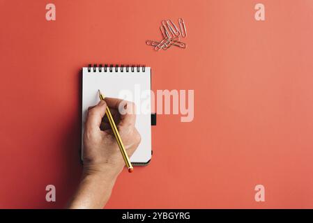 Lay flat of woman hand with pencil writing on blank page of notebook and paperclips on bubble-gum pink background. Conceptual, planning Stock Photo