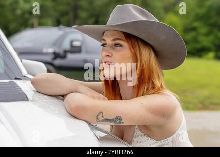 A beautiful redhead model poses in a country setting wearing cowgirl attire against her truck Stock Photo