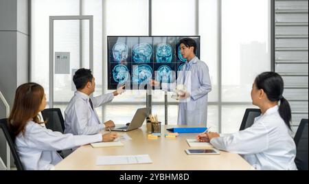 Young medical professional standing and presenting, holding a model of human skull and referencing image of brain scan displayed on a screen. The audi Stock Photo