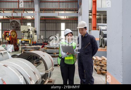 Two people are working in a factory. They are discussing plans while looking at a laptop. They wear safety helmets. Stock Photo