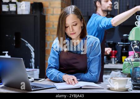 Young woman coffee shop worker in apron using laptop computer at bar counter Stock Photo
