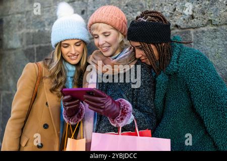 Group of diverse women friends looking at smartphone while shopping in winter. Multicultural friendship, urban lifestyle, and technology use during ho Stock Photo