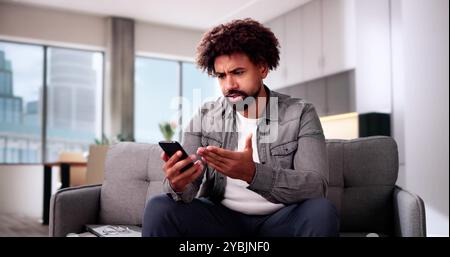 Worried Young Man Looking At Smartphone And Sitting In Front Of Sofa At Home Stock Photo