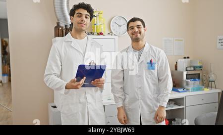 Two men in a laboratory wearing lab coats, one holding a clipboard, embody professionalism and teamwork. Stock Photo
