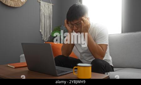 Young man working at home in a living room, appearing stressed while looking at his laptop, surrounded by a cozy interior with a book, coffee cup, and Stock Photo