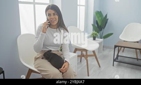 A young woman talks on a phone while holding a chihuahua in a modern veterinary clinic's waiting room. Stock Photo