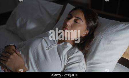 Young woman sleeping peacefully in bed, with brunette hair, wearing white top in bedroom at home, showcasing calm indoor scene of relaxation Stock Photo