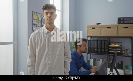 Two hispanic men working in an office with one standing near a window while the other is seated at a desk in front of a computer Stock Photo